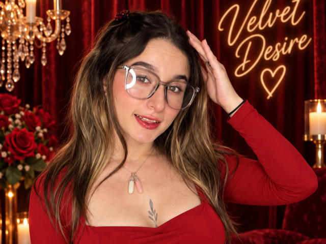 MilaGonz with glasses, red dress, and dog tags necklace, hand in hair, smiling, wavy brown hair, small tattoo on chest, red lipstick, chandelier, candles, roses, and heart.