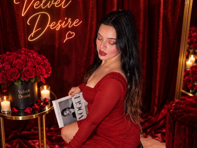 ViolettaRouse with long black hair, wearing a red dress, sitting, holding a photo, surrounded by red roses, candles, and velvet. She has red lipstick and a seductive look.