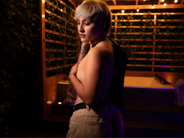 DannyRouds with short blonde hair, topless, covering her tits with arms, wearing high-waisted beige pants, tattooed arm, standing in dimly lit wooden area with string lights.
