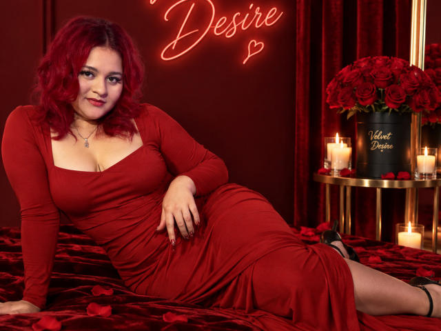 RosalieVittoria with red curly hair, wearing a red dress, showing her big tits, sitting on red velvet, surrounded by candles and roses, smiling at the camera.