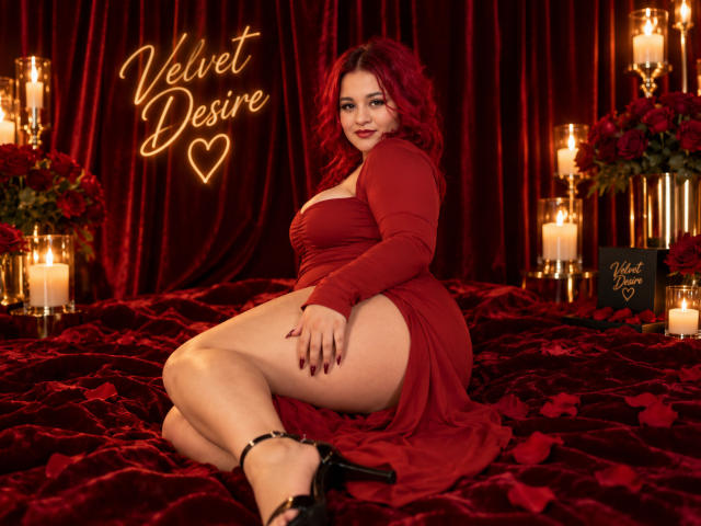RosalieVittoria with red curly hair, red dress, showing her ass, sitting on red velvet, surrounded by candles, wearing black high heels, looking at the camera, smiling.