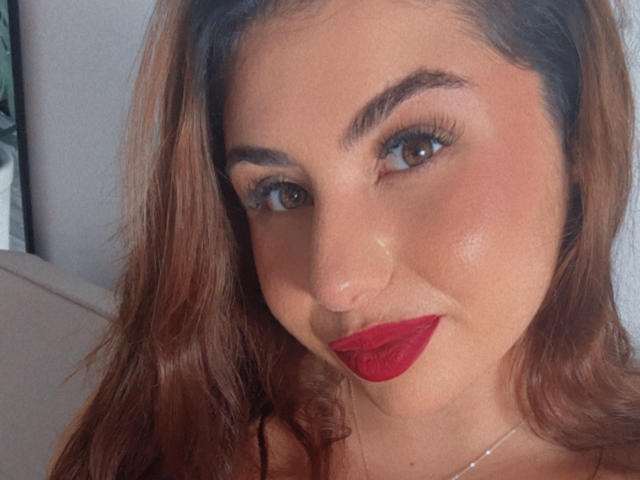 VenusxMusa with red lipstick, brown wavy hair, and subtle makeup, smiling at the camera. She's got a cute nose and brown eyes, looking sexy and attractive.
