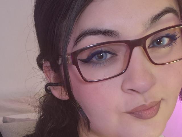 LindaSophieX has fair skin, black hair in a ponytail, brown glasses, blue eyeliner, and pink lipstick, with a slight smirk, looking into the camera.