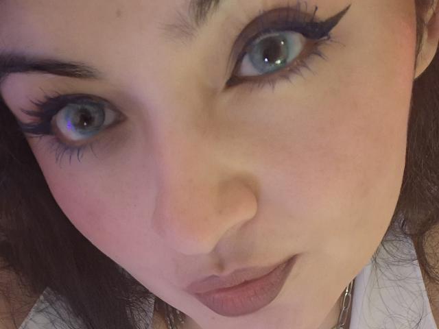 Le visage de LindaSophieX, avec ses yeux bleus, ses longs cils noirs et sa peau rosée. Elle a les cheveux bruns et ondulés, des lèvres roses et pleines, et un léger blush rose sur les joues..