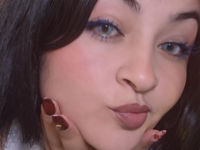 LindaSophieX, avec ses lèvres boudeuses, son eye-liner bleu et ses ongles foncés, se tient la joue, mettant en valeur son joli visage et son ongle doré brillant, avec un blush rose pâle sur sa peau..
