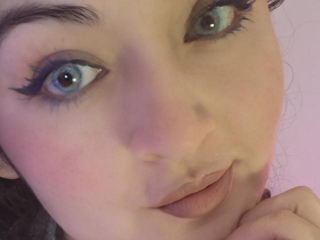 Le visage de LindaSophieX, avec ses yeux bleus soulignés d'un trait de khôl noir épais, ses lèvres rosées légèrement entrouvertes, sa main près du menton, un blush rose pâle sur les joues, une peau claire, des sourcils foncés, et un léger sourire..