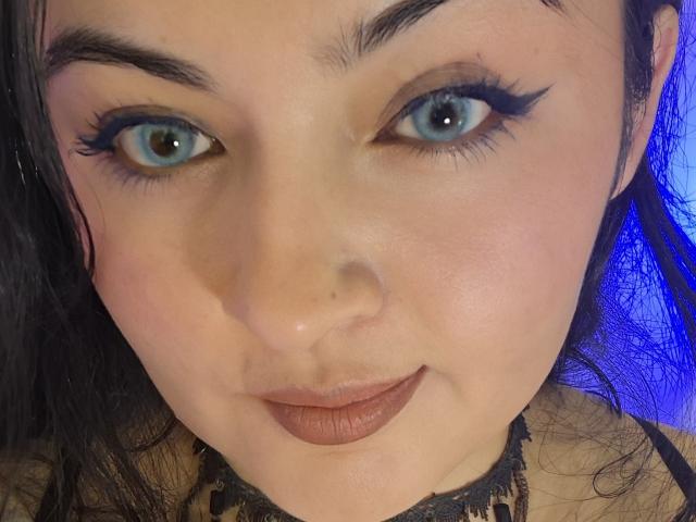 Le visage de LindaSophieX, avec ses yeux bleus, son eye-liner noir et son rouge à lèvres brun, portant un collier en dentelle, affichant un léger sourire, et ses cheveux noirs sont partiellement visibles..
