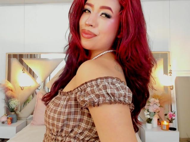 ElizaNior with red hair, plaid off-shoulder top, pearl necklace, and pouty lips, showing her tits slightly, smiling at the camera.