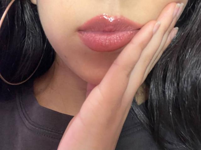 HannaThorne's glossy pink lips, hand gently touching them, wearing large gold hoop earrings, dark wavy hair, in a black top, with manicured nails and a sexy, sultry look.