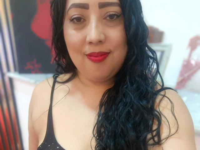 AmberMoretti has curly black hair, thick eyebrows, red lipstick, and is wearing a black spaghetti-strap top. Her medium-sized tits are slightly visible. She's smiling at the camera.