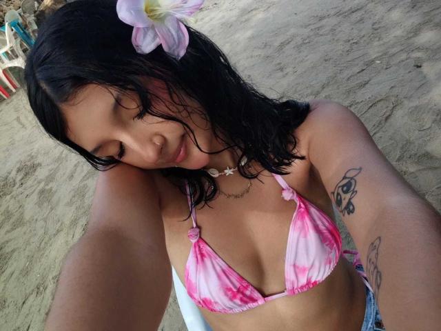 PaulineRey with wet black hair, pink tie-dye bikini top, flower in hair, tattoo on arm, smiling, selfie, tan skin, small tits, necklace, sitting on sandy beach.
