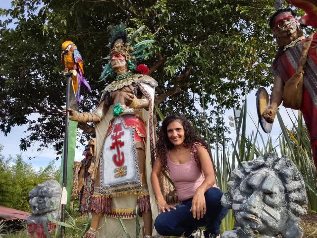 SnowLily with curly brown hair, pink tank top, and blue jeans, kneeling in front of a tall, colorful, Native American-style statue with a bird and another similar statue on the right.