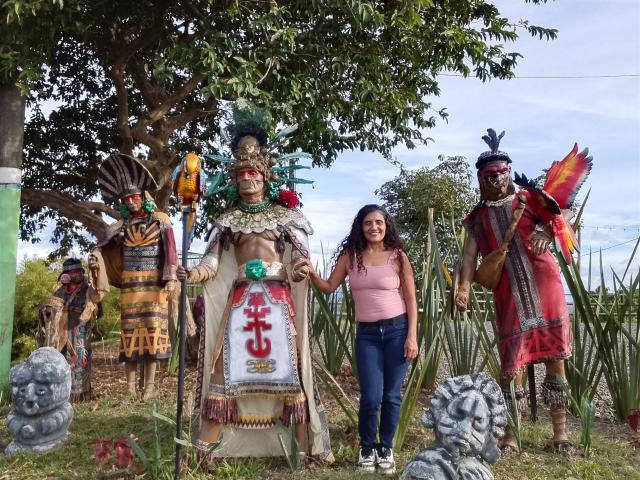SnowLily stands with four tall, colorful wooden statues wearing headdresses and traditional clothing, featuring large breasts and prominent penises. She's in a pink top, holding a statue's hand.