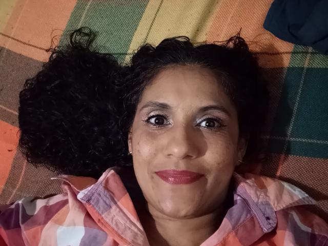 SnowLily's face, lying down with curly black hair, wearing a pink and purple plaid shirt, has brown eyes, pink lipstick, and a slight smile, looking up at the camera.