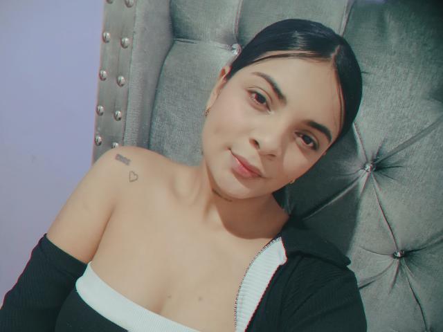 KarynNancy, con capelli scuri, pelle chiara, seno piccolo, top nero con spalline scoperte, poltrona grigia con decorazioni, tatuaggio sulla spalla destra, sorridente, labbra rosa chiaro, trucco leggero, espressione rilassata..