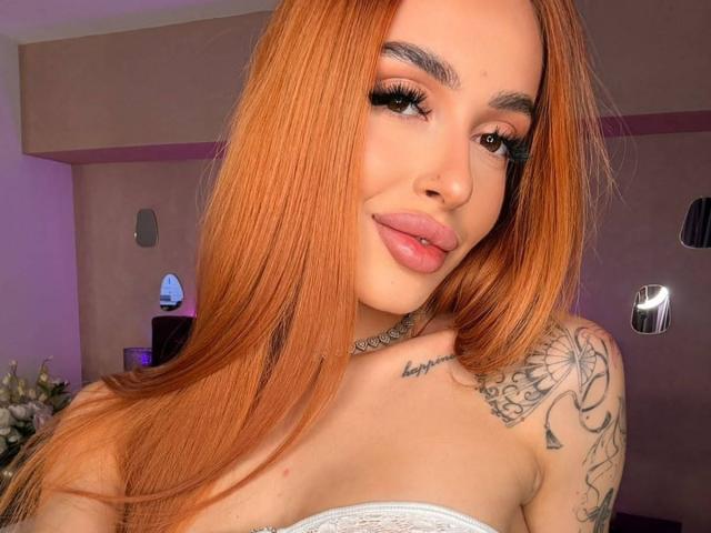 MarissaFex has long red hair, full pink lips, heavy eyelashes, and tattoos on her shoulder and chest. Her white strapless top reveals her tits. She's smiling slightly.