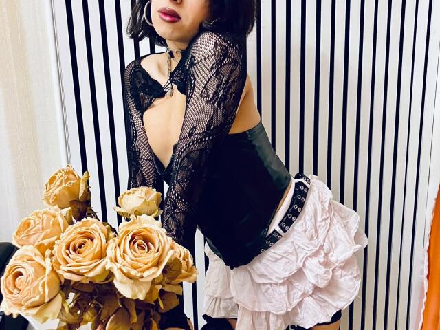 FemCupid with black lace top, low-cut revealing ass, black corset, and pink ruffled skirt, leaning forward, dark hair, red lipstick, holding beige roses, striped background.