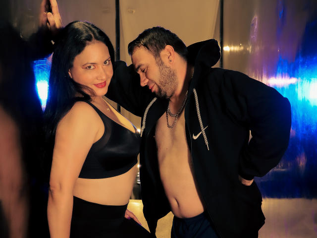 MarihamMurath with black bra and high-waisted black pants, standing next to a shirtless man in a black hoodie, leaning in with a playful smile. Her tits are visible.