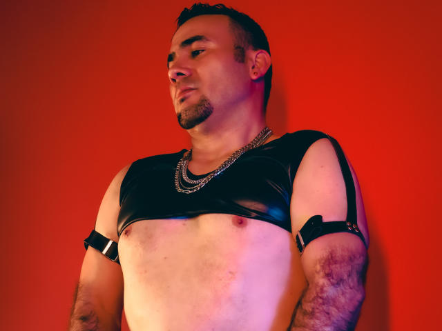 MarihamMurath with short dark hair, beard, and chain necklace, wearing a black leather chest harness, exposing his nipples and hairy arms, against a red background.