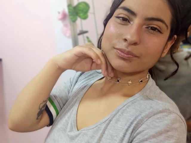 AngelAlba with a cute face, light brown skin, and dark hair, wearing a gray V-neck shirt, gold necklace, and a green and black tattoo on her right arm.