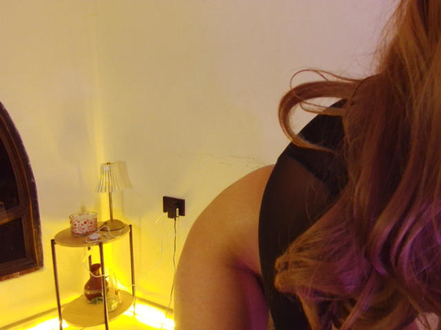 Ilaynna's butt in black sheer lingerie, her wavy brown hair in the foreground, yellow lamp on a small table with a teacup and vase, and a black plug.