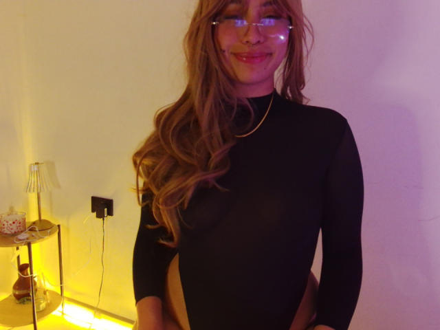 Ilaynna with long brown hair, glasses, and a black long-sleeve top, showing her small tits and a visible ass, standing in a room with yellow lighting.