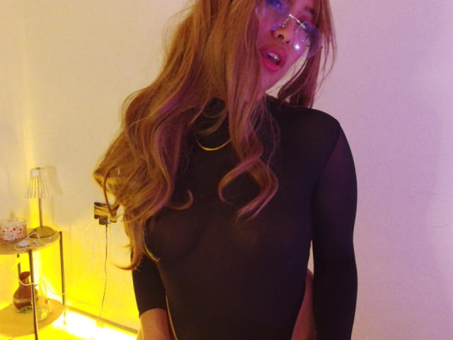 Ilaynna with long red hair, wearing a sheer black top, showing her small tits, and clear glasses, standing in a room with yellow lighting.