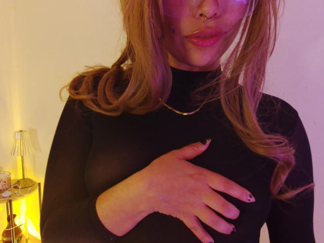 Ilaynna with pigtails, nose ring, and black sheer top, hand covering her tits, pinkish-purple lighting, gold necklace, and black nail polish, smiling slightly.