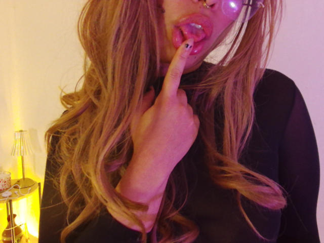 Ilaynna with long blonde hair, black top, and glasses, sticking her finger into her shiny pink lips, showing black nail polish, with a yellow lamp in the background.