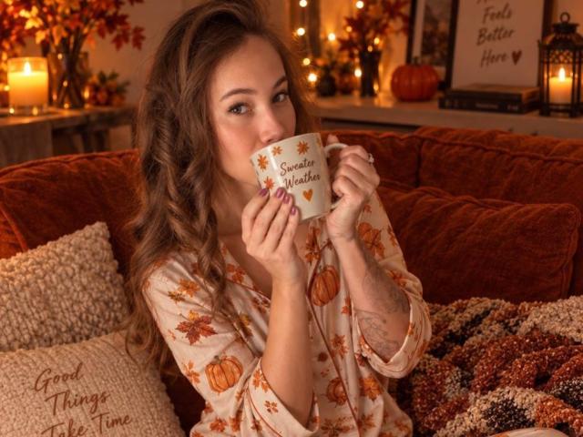 AmmaLoyXX with wavy brown hair, sipping from a pumpkin-themed mug, wearing a fall-patterned shirt, sitting on a red couch with cozy blankets. Warm, autumnal, and cute.