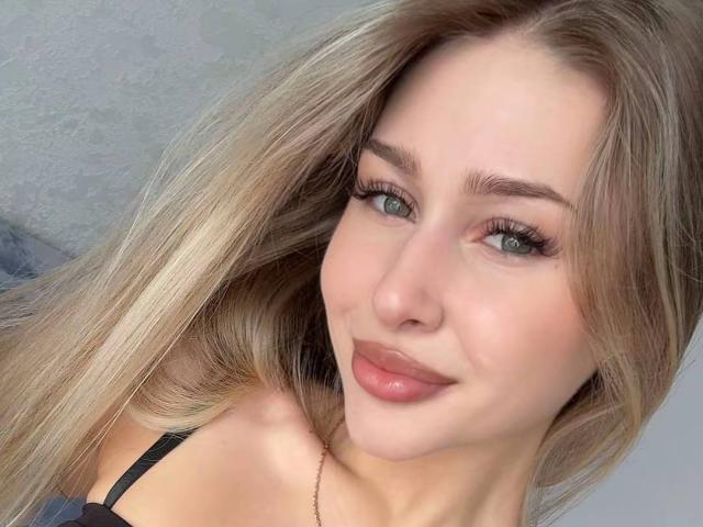 MilagrosEdmisten has long blonde hair, blue eyes, and pink lips with a subtle smile. She's wearing a black strap, showing her bare shoulders. Her tits are not visible.