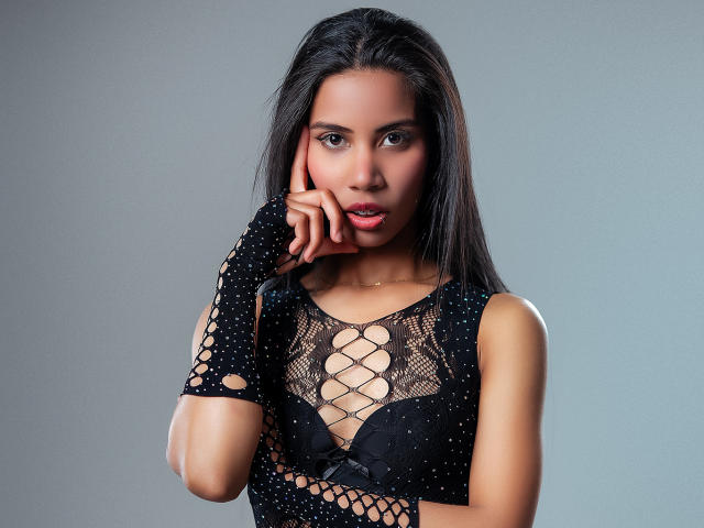 LauraJackson with long black hair, wearing a black, lace-up, fishnet top, exposing her boobs, touching her chin, wearing a fingerless, black, polka-dot glove, gray background.