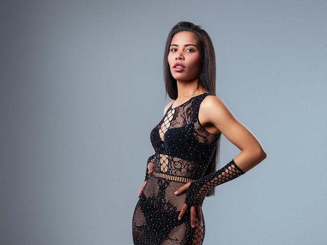 LauraJackson with long black hair, wearing a black lace dress with mesh, showing her tits and pussy through the material, and lace finger gloves, standing with hands on hips.
