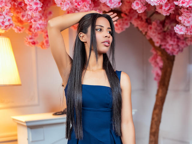 LauraJackson with long black hair, blue dress, arm raised, looking to the side, pink cherry blossoms in background, warm light from lamp on left, gold necklace.
