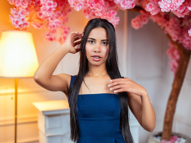 LauraJackson with long black hair, wearing a blue dress, has her hands near her head, standing in front of pink cherry blossoms, under warm indoor lighting.