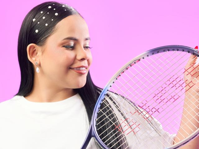 NixieRose with dark hair, white shirt, and pearl earrings, holding a tennis racket, smiling at it. Pink background, white polka dot headband, visible arm with black sleeve.