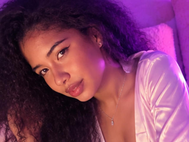 Tifannyy has curly black hair, brown eyes, and a slight pout. She's wearing a shiny white top, and a silver necklace with a small pendant. Pink and purple light.