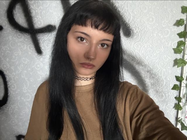 IrisNetti, met lang zwart haar, bruine ogen en een neusring, draagt een bruin topje en een zwarte choker, heeft een serieuze uitdrukking, en een wijnrank is gedeeltelijk zichtbaar..