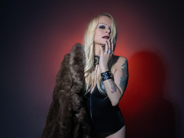 Immerlina, con cabello rubio, top de cuero negro, abrigo de piel marrón, tatuajes, lápiz labial oscuro y pulseras con púas, tocándose la barbilla, con una mirada seductora, mostrando sus senos. Fondo de luz roja..