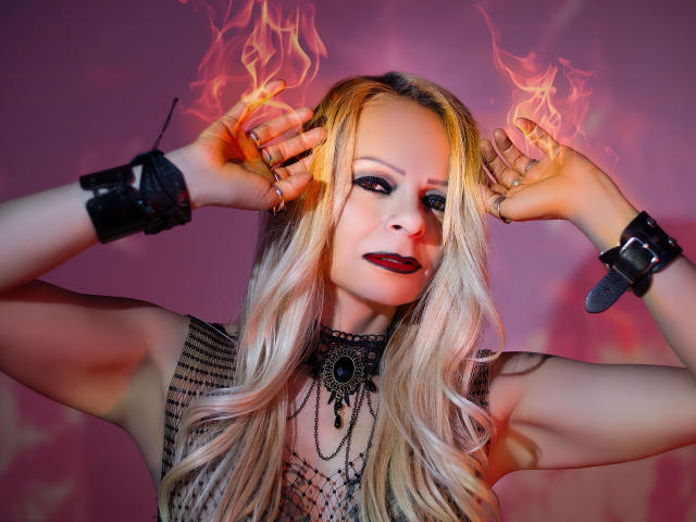 Immerlina met blond haar, met vuur boven haar hoofd, draagt een zwarte ketting, rode lippenstift en zwarte manchetten, met armen omhoog, een verleidelijke blik, een vurige oranje en roze gloed..