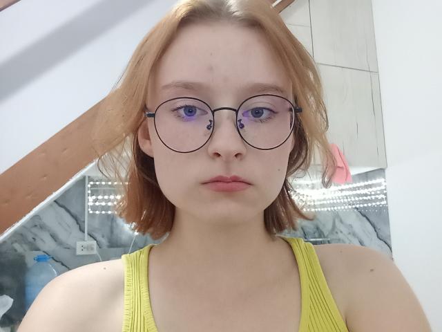 GeorgianaLefleur has pale skin, light brown shoulder-length hair, round glasses, and wears a yellow tank top, showing her small tits and a pimple on her forehead.