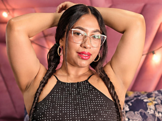 SoftLatina with braided hair, red lipstick, and glasses, arms raised, wearing a black, dotted, sleeveless top, showing her tits, standing against a pink, tufted headboard.