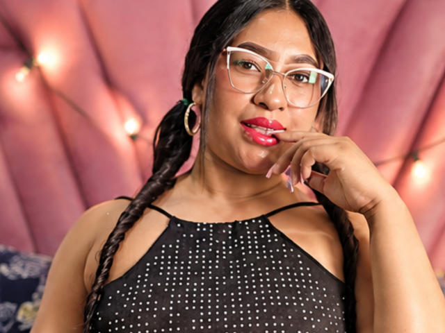 SoftLatina with braided hair, wearing glasses, red lipstick, and a black, sparkly top, touching her chin with a finger, hoop earrings, and a confident, sexy look.