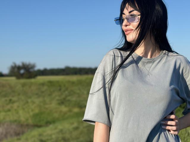 Xioni has long black hair, wears glasses, and a loose gray t-shirt, standing with a hand on her hip, smiling slightly, with clear blue sky and green field behind.
