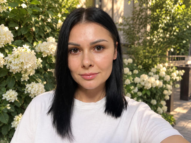 Ariana Diaz mit schwarzem, glattem Haar, braunen Augen und heller Haut, trägt ein weißes T-Shirt, hat ein leichtes Lächeln und zarte, rosafarbene Lippen und steht vor weißen Blumen..