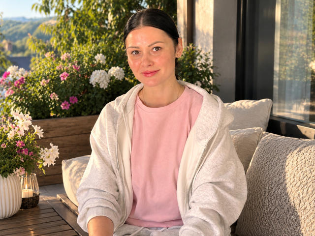 Ariana Diaz mit heller Haut, einem rosafarbenen Hemd und einer grauen Kapuzenjacke, sitzt draußen, mit schwarzem Haar im Pferdeschwanz, einer kleinen Nase, einem süßen Lächeln und einem koketten Blick, umgeben von Blumen und Kissen..