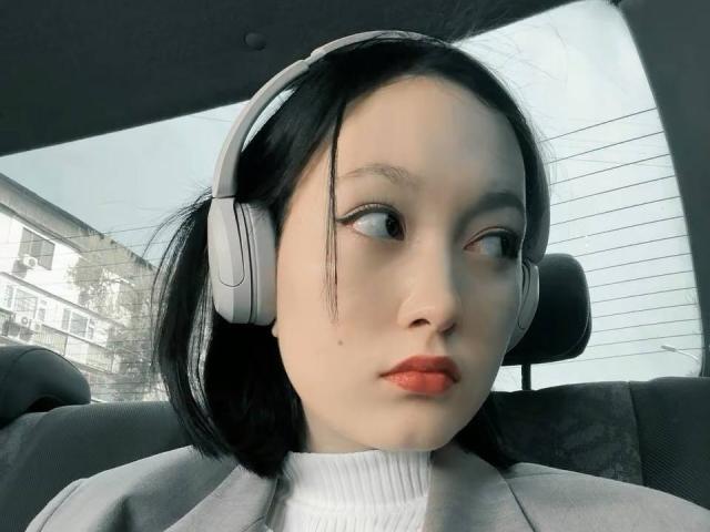 LoidaBoehl, with pale skin and black hair, wearing grey headphones, has a small mole on her left cheek, and red lipstick, looking sideways in a car.