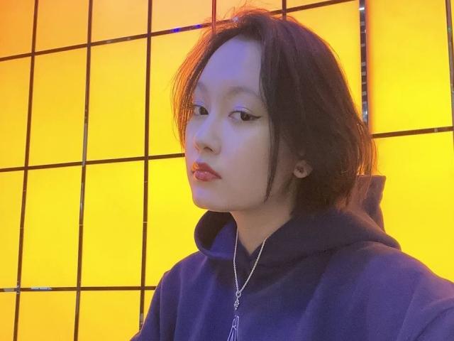 LoidaBoehl with pale skin, black eyeliner, and pink lips, wearing a blue hoodie and silver necklace, standing against a yellow grid wall. Her black hair is slightly messy.