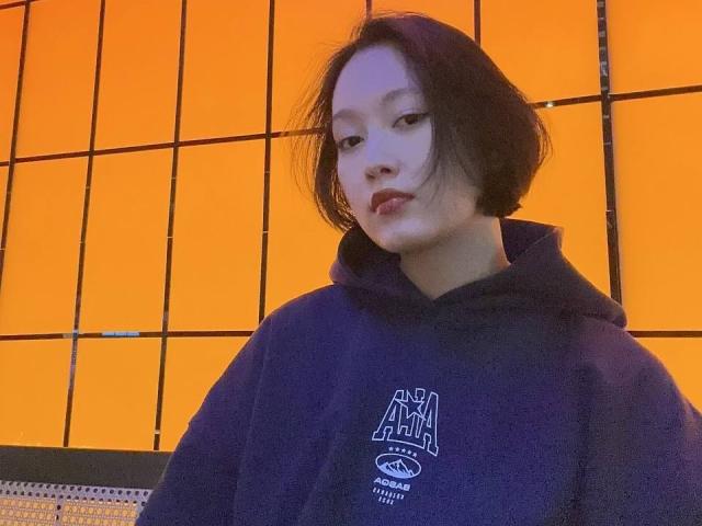 LoidaBoehl with short black hair, wearing a dark blue hoodie, has a serious expression, orange-lit background, and a subtle smile with red lipstick. Her face is slightly blurry.