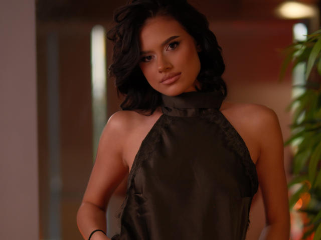 IvyVeil with wavy black hair, wearing a dark, shiny, halter-style top, showing bare shoulders and a confident, sultry expression. Her tits are partially visible, looking sexy.